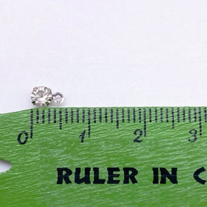 May include: A small, clear rhinestone with a silver loop attached. The rhinestone is about 0.25 inches in diameter. A ruler is in the background to show the size.