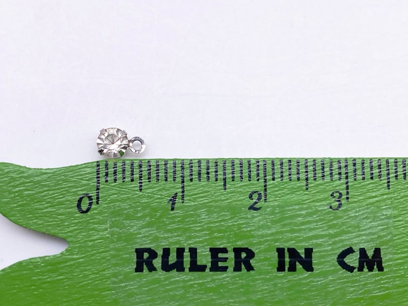 May include: A small, clear rhinestone with a silver loop attached. The rhinestone is about 0.25 inches in diameter. A ruler is in the background to show the size.