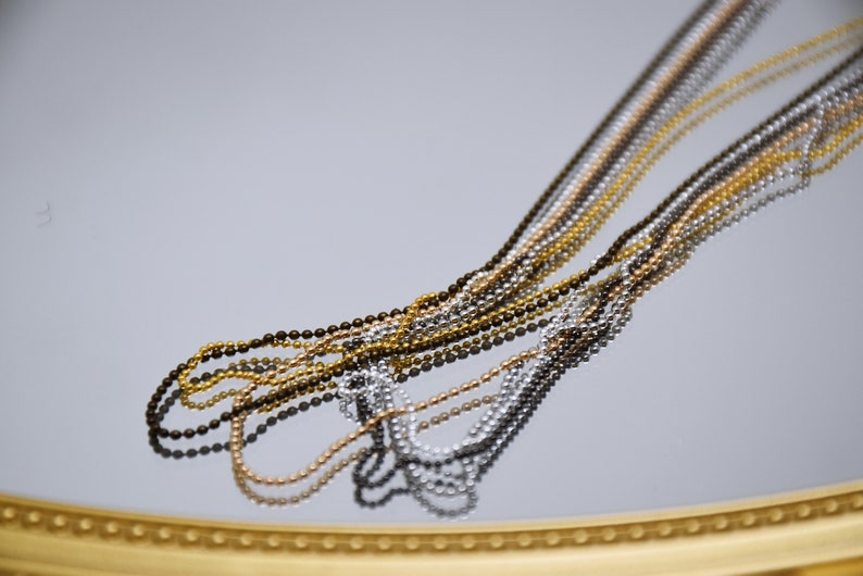 May include: A close-up of multiple chains of different colors and metals. The chains are made of small, round beads and are arranged in a fan shape. The chains are reflected in a mirror surface.