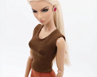 May include: A fashion doll with long blonde hair, wearing a brown sleeveless knit top and rust-colored shorts. The doll features articulated joints, dark eye makeup, and a single visible earring with a bright pink stone set in a silver frame.