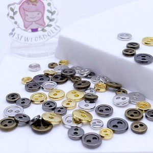 Puede incluir: Una colección de pequeños botones de dos agujeros en tonos plateado, dorado y bronce. Los botones están esparcidos sobre una superficie blanca.