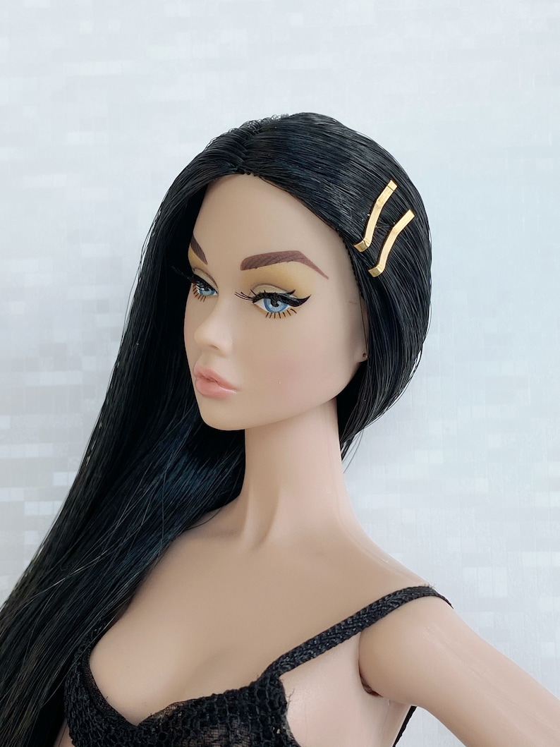 May include: A doll with long black hair wearing a black top and gold hair clips.