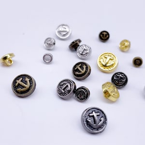 Może przedstawiać: Zestaw metalowych guzików z motywem kotwicy. Guziki są w różnych kolorach, w tym złote, srebrne i czarne.