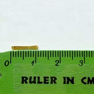 May include: A gold-coloured metal bar measuring approximately 1 centimetre in length, placed on a green ruler with black markings.