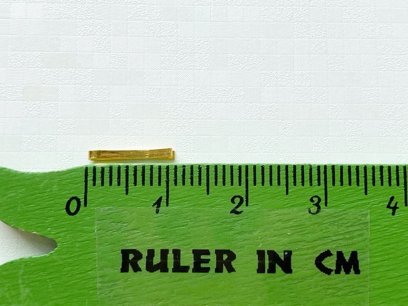 May include: A gold-coloured metal bar measuring approximately 1 centimetre in length, placed on a green ruler with black markings.