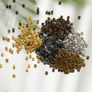 May include: A close-up of a variety of small metal buttons in gold, silver, and bronze colours. The buttons are scattered on a white surface.