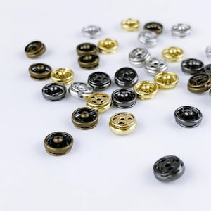 Puede incluir: Un conjunto de broches metálicos de varios colores, incluyendo dorado, plateado, negro y bronce. Los broches redondos tienen un agujero central y una base con púas, dispersos sobre una superficie blanca. Probablemente se usan para proyectos de costura.