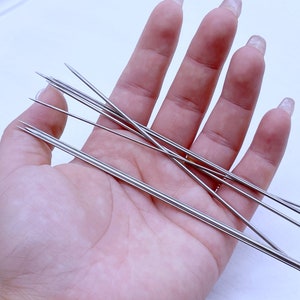 May include: A set of eight silver beading needles, held in a hand, against a white background.