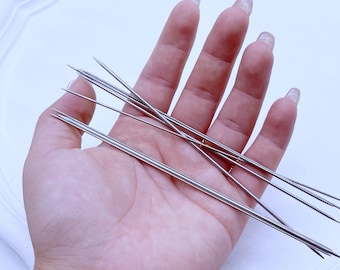 May include: A set of eight silver beading needles, held in a hand, against a white background.