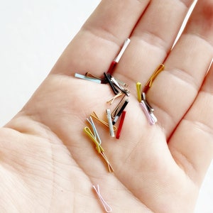 May include: A hand holding a selection of small, colourful hair clips. The clips are made of metal and plastic and come in a range of colours, including gold, silver, black, red, and blue.