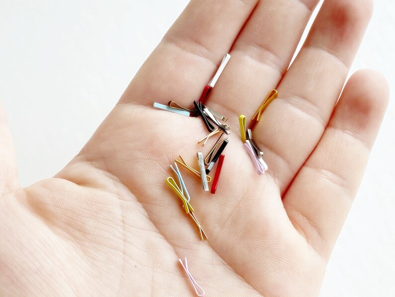 May include: A hand holding a selection of small, colourful hair clips. The clips are made of metal and plastic and come in a range of colours, including gold, silver, black, red, and blue.