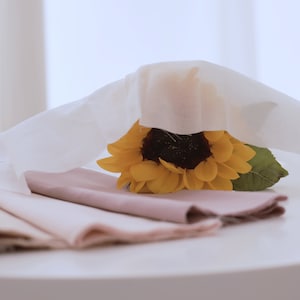 May include: A stack of four pale pink fabric squares with a yellow sunflower on top, partially covered by a white sheer fabric.