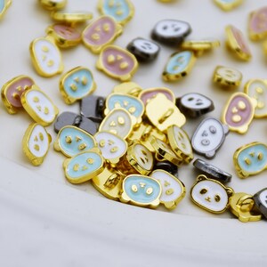 Puede incluir: Una colección de pequeños botones ovalados con caras de panda. Los botones son de color dorado con detalles en esmalte blanco, azul y rosa.