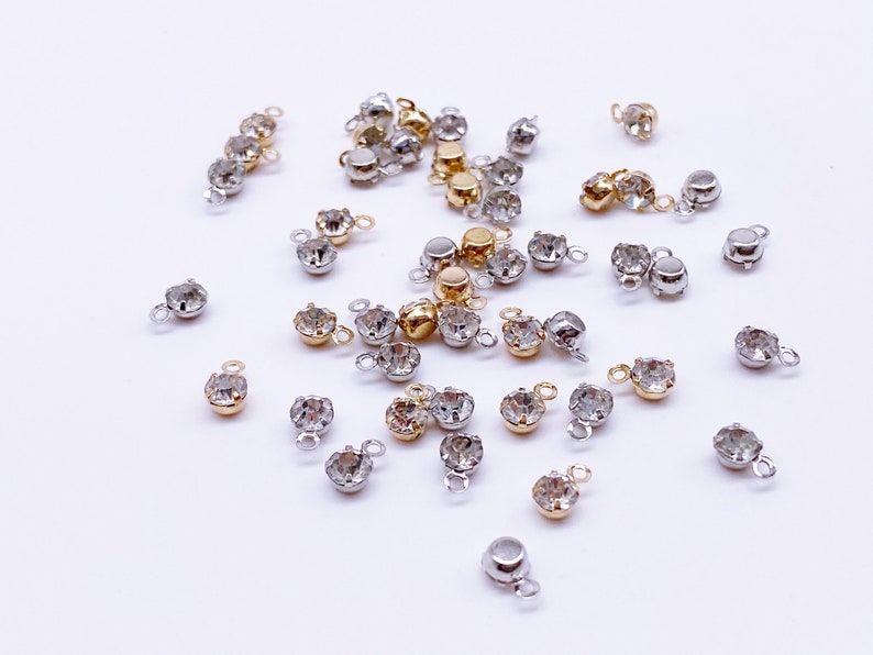 May include: A collection of clear crystal rhinestones with gold and silver settings. Each rhinestone has a small loop on the back for attaching to jewelry or other crafts.