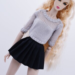 May include: A doll wearing a gray knitted sweater with a black pleated skirt. The doll has long blonde hair and is wearing red earrings.