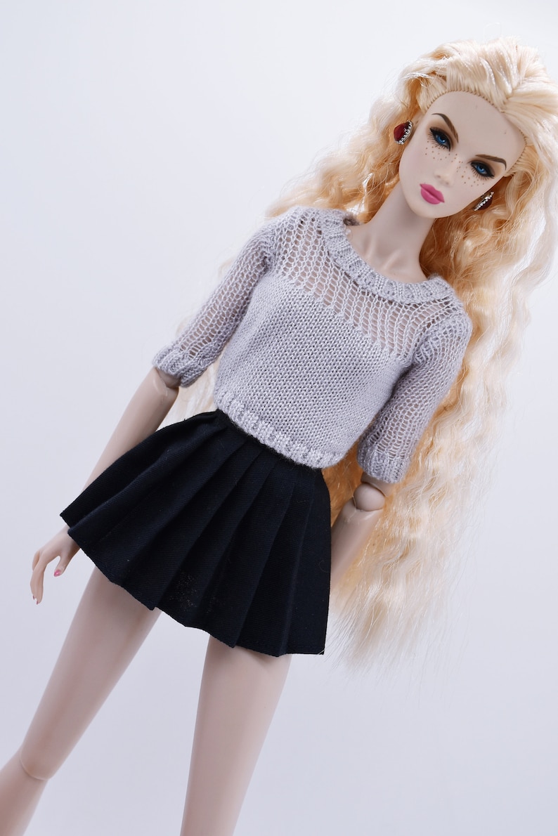May include: A doll wearing a gray knitted sweater with a black pleated skirt. The doll has long blonde hair and is wearing red earrings.