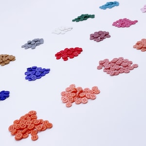 May include: A collection of small, round buttons in various colors, including red, blue, pink, and brown, arranged on a white surface. Each button has four holes and a slightly raised edge. The buttons are evenly spaced, creating a visually appealing pattern.
