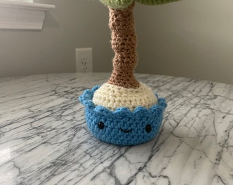 May include: A crocheted palm tree with a green crown and brown trunk, standing on a blue and white crocheted island with a smiling face.