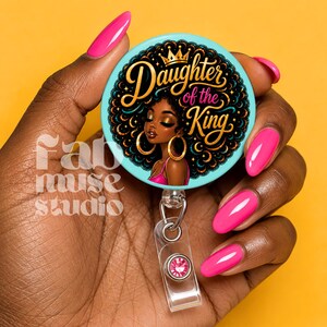 May include: A round badge reel with the text "Daughter of the King" and an illustration of a Black woman with gold hoop earrings. The badge reel has a light blue background and a clear plastic retractable cord with a pink gem.
