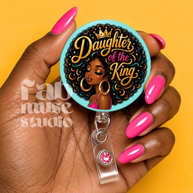 May include: A round badge reel with the text "Daughter of the King" and an illustration of a Black woman with gold hoop earrings. The badge reel has a light blue background and a clear plastic retractable cord with a pink gem.