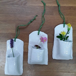 May include: Three white ceramic wall planters with green twine hanging loops. Each planter has a small pocket for holding a single flower. The planters are shown with flowers in them: lavender, pink begonia, and yellow jasmine.