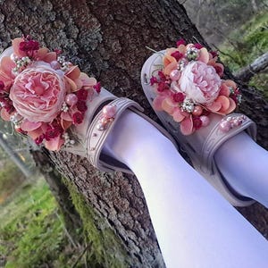 May include: A pair of light gray clogs adorned with a floral arrangement of pink and peach-colored flowers, berries, and pearls. The clogs are decorated with pink pearls on the straps. The background shows a tree trunk and greenery.