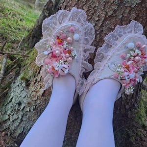 May include: A pair of beige slippers adorned with pink floral embellishments, pearls, and lace trim. The slippers are decorated with pink and white flowers and are set against a natural background.