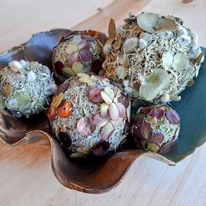 May include: An arrangement of moss-covered spheres decorated with dried flowers and foliage, presented in a rustic, organic-shaped ceramic bowl. The spheres vary in size and are accented with shades of purple, orange, and green.