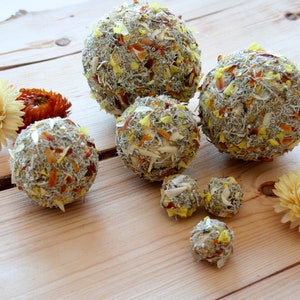May include: A collection of dried flower balls in various sizes, ranging from small to large, with a mix of white, yellow, and brown petals. The balls are arranged on a wooden surface.