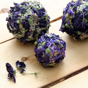 Puede incluir: Tres objetos esféricos cubiertos de pétalos de flores secas moradas y verdes descansan sobre una superficie de madera clara. Unos pocos pétalos sueltos están esparcidos cerca. Los objetos parecen decorativos o para manualidades.