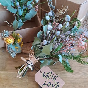 May include: A collection of dried flower bouquets with a variety of textures and colors, including green eucalyptus leaves, pink and white flowers, and fluffy gray accents. Each bouquet is tied with twine and includes a tag that says "With Love".