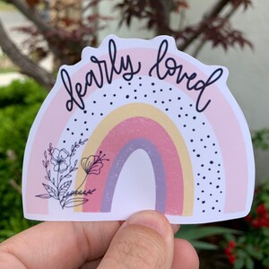 May include: A pink and yellow rainbow sticker with black polka dots and the text "dearly loved" in black script. A black line drawing of a flower is in the bottom left corner.