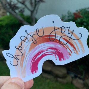May include: A sticker with a rainbow design in shades of pink, orange, and brown. The rainbow has a white outline and the words "choose joy" are written in black cursive.