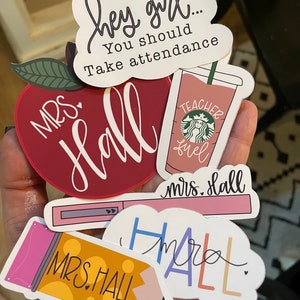 May include: A set of six stickers featuring the name "Mrs. Hall" in various styles. The stickers include a red apple, a coffee cup, a pencil, a lipstick, and two cloud-shaped stickers.