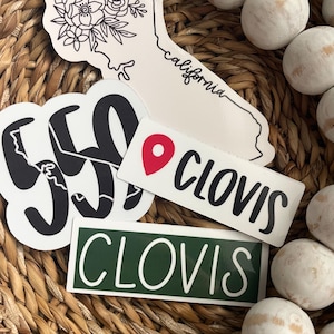 May include: A collection of stickers featuring the words "Clovis" and "California" with a black and white floral design. The stickers are on a woven wicker surface.