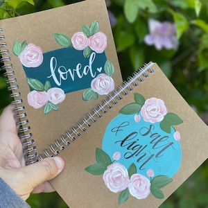 May include: Two brown spiral bound notebooks with painted floral designs. The first notebook has a green background with the word "loved" in white script. The second notebook has a turquoise circle with the words "Salt & Light" in black script.