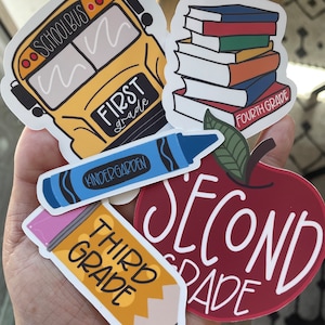 May include: A set of five colorful stickers featuring school-themed designs. The stickers include a yellow school bus with the text "School Bus" and "First Grade", a stack of books with the text "Fourth Grade", a blue crayon with the text "Kindergarten", a red apple with the text "Second Grade", and a yellow pencil with the text "Third Grade".