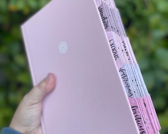 May include: A light pink book with labeled tabs. The tabs are white with pink edges and feature handwritten text. The book has a white ribbon bookmark and a small floral design on the cover. The book is held against a blurred green background.