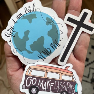 May include: A collection of stickers featuring religious themes. One sticker shows a blue and green globe with the words "here I am Lord, send me." Another is a black cross on a white background. The third sticker depicts a van with the words "Go Make Disciples."
