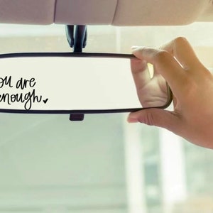 May include: A car's rearview mirror with the words "you are enough" in black script. A hand is adjusting the mirror. The image has a soft, diffused lighting, suggesting a calm and reflective mood.