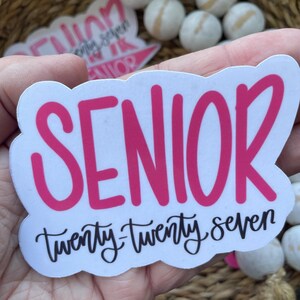 May include: A white sticker with a pink "SENIOR" text and "twenty-twenty seven" in black script. The sticker has a cloud-like border. Other stickers and white wooden beads are in the background.
