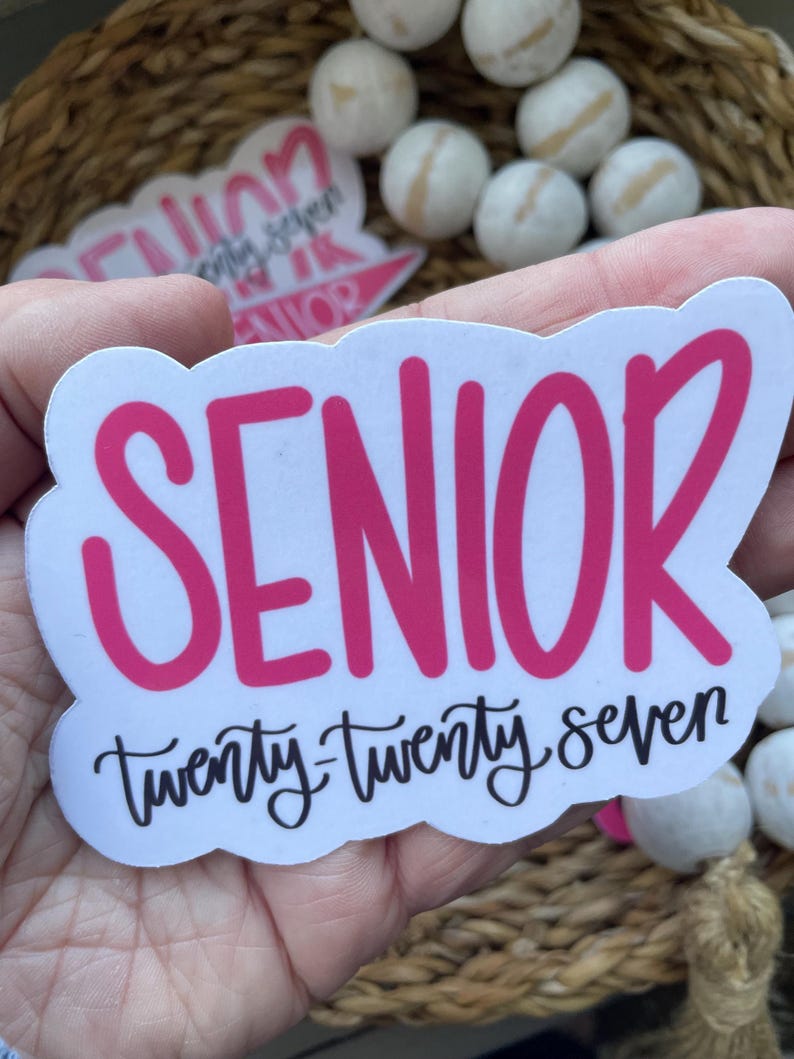 May include: A white sticker with a pink "SENIOR" text and "twenty-twenty seven" in black script. The sticker has a cloud-like border. Other stickers and white wooden beads are in the background.
