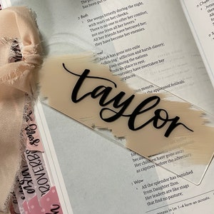 May include: A close-up of a beige and brown acrylic name card with the name "Taylor" written in black cursive lettering. The card is sitting on top of an open book with text about the book of Lamentations in the Bible.