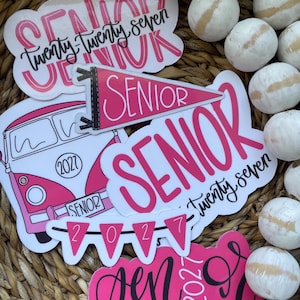 May include: A collection of pink and white stickers with the word "Senior" and the year "2027" in various fonts. One sticker features a pink and white bus. The stickers are arranged in a woven basket with decorative white balls.