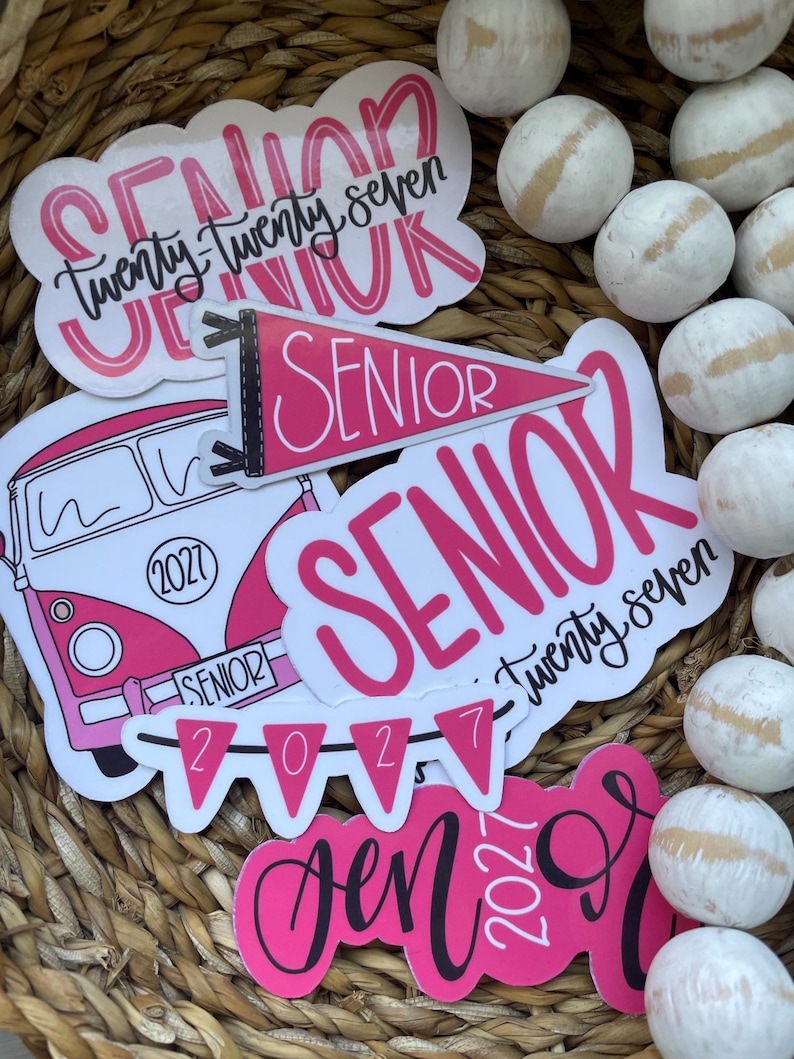 May include: A collection of pink and white stickers with the word "Senior" and the year "2027" in various fonts. One sticker features a pink and white bus. The stickers are arranged in a woven basket with decorative white balls.
