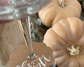 May include: A brown paper place card with a white handwritten name "christi" attached to a clear wine glass. The glass is sitting on a wooden surface with two small pumpkins.