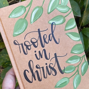 May include: A brown notebook with green painted leaves and the text "rooted in christ" in black calligraphy.