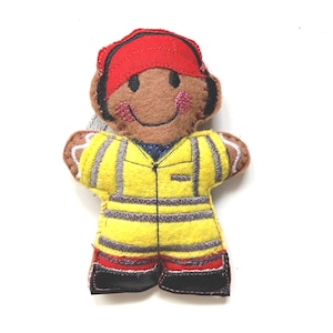 May include: A felt firefighter ornament. The ornament is shaped like a person with a brown face, a red helmet, and a yellow uniform with reflective stripes. The uniform has red and black accents. The ornament has a smiling face.
