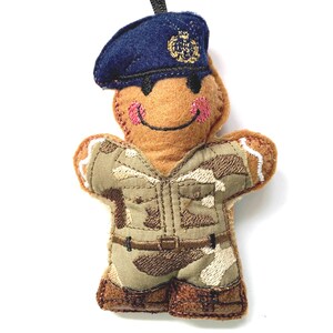 May include: A gingerbread person wearing a camouflage uniform and a blue hat with a gold emblem. The gingerbread person has a smiling face with pink cheeks.