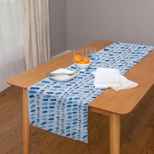 May include: A blue and white table runner with an abstract brushstroke pattern. The runner is laid on a wooden table with a white bowl of oranges and two clear glasses.
